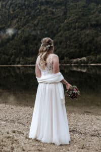 Eine Frau in einem weißen Hochzeitskleid steht an einem ruhigen See, umgeben von üppigem grünen Wald. Sie steht mit dem Rücken zur Kamera und hält einen Strauß mit roten Blumen in der Hand. Als Teil ihrer Hochzeit ist ihr Haar mit einer Blumenkrone geschmückt und ein Schal liegt elegant über ihren Schultern.
