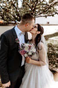 Ein Paar küsst sich an seinem Hochzeitstag im Freien. Der Bräutigam, in dunklem Anzug und Fliege, umarmt seine Braut in weißem Kleid und Schleier. Sie hält einen Strauß weißer und violetter Blumen in den Händen, während sie unter einem Baum stehen. Ihr perfekter Tag symbolisiert die Freude des Heiratens.