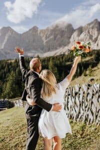 Ein Paar in festlicher Kleidung steht auf einem grasbewachsenen Hügel und feiert seine Hochzeit vor der Kulisse majestätischer Berge. Die Frau hält einen Strauß orangefarbener und weißer Blumen und streckt freudig die Arme in die Höhe. Ein Holzzaun und ein bewaldeter Hügel vervollständigen die malerische Szene unter einem klaren blauen Himmel.