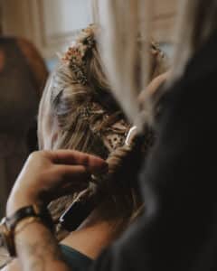 Eine Person frisiert das Haar einer Frau und lockt es mit einem Werkzeug für ihre Hochzeit. Das Haar ist mit kleinen Blumen geschmückt, perfekt für diesen besonderen Anlass. Der Fokus liegt auf den komplizierten Details der Frisur und der Accessoires und fängt die Essenz einer sorgfältigen Hochzeitsplanung ein.