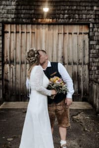 Ein Paar küsst sich vor einem hölzernen Scheunentor und verkörpert damit die Essenz ihrer Traumhochzeit. Die Braut in einem langärmeligen Spitzenkleid mit einem Blumenstrauß in der Hand und der Bräutigam in einer Weste und traditionellen Ledershorts stehen auf nassem Bürgersteig unter einer leuchtenden Außenlampe.