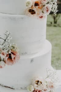 Eine dreistöckige weiße Hochzeitstorte, geschmückt mit blassrosa und weißen Rosen, Schleierkraut und Grünpflanzen. Der Hintergrund ist verschwommen und suggeriert eine ideale Umgebung im Freien für Hochzeiten.