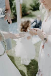 Eine Nahaufnahme von zwei Frauen, die bei einer Veranstaltung im Freien, möglicherweise einer Hochzeit, Händchen halten. Eine trägt einen Ring, die andere eine Uhr. Im unscharfen Hintergrund sitzen ein Kind in einem weißen Kleid und ein Erwachsener im Gras. Die Szene ist in helles Tageslicht gehüllt.