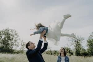Auf einer Wiese unter klarem Himmel hebt ein Mann freudig ein Kind in die Luft. Beide lächeln breit. Ein junges Mädchen steht daneben und grinst ebenfalls. Diese freudige Szene erinnert an Erinnerungen, die bei Hochzeiten gemacht wurden – vielleicht während einer Hochzeit oder bei sorgfältiger Hochzeitsplanung.