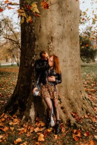 Ein Paar umarmt sich vor einem großen Baumstamm, umgeben von Herbstlaub. Die Frau in einem Rock und Stiefeln mit Leopardenmuster und der Mann in einer Lederjacke strahlen Wärme aus, während sie in dieser gemütlichen Herbstkulisse von ihrer bevorstehenden Hochzeit träumen.