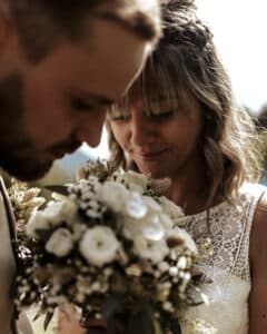 Braut und Bräutigam berühren sanft ihre Stirnen und lächeln beide sanft an ihrem Hochzeitstag. Die Braut hält einen Strauß weißer und blasser Blumen. Sie sind draußen, mit einem leicht verschwommenen Hintergrund, der einen sonnigen Tag suggeriert und die Freude einer perfekten Hochzeitsplanung verkörpert.