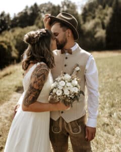 Eine Braut im weißen Kleid und ein Bräutigam in traditioneller Kleidung teilen einen intimen Moment im Freien, während sie ihre Hochzeit feiern. Die Braut hält einen Strauß weißer Blumen und der Bräutigam trägt einen Filzhut. Sie stehen auf einem grasbewachsenen Weg mit Bäumen im Hintergrund und verkörpern perfekte Hochzeitsplanung.