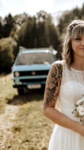 Eine Braut in einem Spitzenhochzeitskleid hält einen Blumenstrauß und steht im Freien mit einem Tattoo auf dem Arm. Unter dem teilweise bewölkten Himmel, inmitten von Bäumen und durchdachter Hochzeitsplanung, parkt im Hintergrund ein blaues Oldtimer.