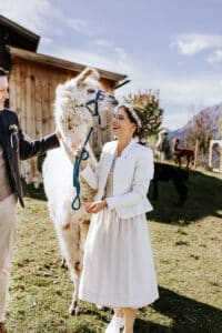 Eine Person in weißem Outfit steht lächelnd vor einem Lama auf einem grasbewachsenen Hügel und denkt vielleicht über Ideen für eine unvergessliche Hochzeit nach. Eine andere Person hält sanft den Kopf des Lamas. Im Hintergrund streifen Alpakas unter dem klaren blauen Himmel in der Nähe eines rustikalen Gebäudes umher.