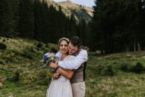 Ein Paar umarmt sich auf einer grasbewachsenen Berglichtung, umgeben von Bäumen. Die Frau, die einen Blumenstrauß hält und ein weißes Kleid trägt, lächelt ihren Verlobten in Weste und Hemd an. Die Szene ist friedlich und romantisch, perfekt für ihre Traumhochzeit mit majestätischen Bergen als Kulisse.