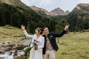 Ein fröhliches Paar im Hochzeitskleid steht in einer malerischen Berglandschaft und feiert seine Hochzeit. Die Braut hält einen Strauß bunter Blumen und beide haben ihre Arme in der Nähe eines fließenden Bachs und eines üppigen grünen Waldes freudig in die Höhe gestreckt.