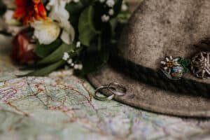 Ein traditioneller Filzhut mit dekorativen Nadeln ruht auf einer detaillierten Karte, begleitet von zwei silbernen Ringen – eine subtile Anspielung auf die Hochzeitsplanung. Ein Strauß leuchtender Blumen ist teilweise im Hintergrund sichtbar und verleiht einen Hauch von Farbe.