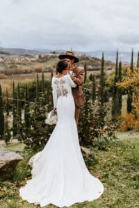 Ein Paar umarmt sich im Freien und verkörpert die Essenz der Hochzeitsplanung. Das lange weiße Kleid der Braut weist aufwendige Rückendetails auf, während der Bräutigam einen braunen Anzug und Hut trägt. Sie stehen auf Gras, mit einer malerischen Hügellandschaft im Hintergrund unter einem bewölkten Himmel, und fangen die Freude ihrer Hochzeit ein.
