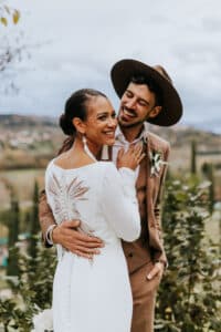 Das lächelnde Paar strahlt an seinem Hochzeitstag vor Freude. Die Frau glänzt in einem weißen Kleid mit aufwendigen Rückendetails, während der Mann sie in einem braunen Anzug und einem breitkrempigen Hut ergänzt. Sie stehen eng beieinander inmitten der malerischen Außenkulisse und verkörpern Liebe und Eleganz.