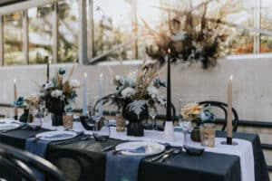 Ein wunderschön gedeckter Esstisch mit schwarzen und weißen Akzenten, perfekt für eine Hochzeit. Er verfügt über Blumenarrangements mit blauen und beigen Blüten, brennende Stabkerzen und elegantes Geschirr. Sonnenlicht strömt durch die großen Fenster im Hintergrund und schafft eine verträumte Atmosphäre für Hochzeiten.
