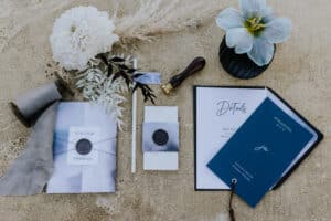 Ein flacher Stapel Hochzeitspapeterie auf Sand enthält eine Blume, eine Spule Band, einen Wachssiegelstempel und Karten. Die Karten zeigen elegante Kalligraphie mit Namen und Veranstaltungsdetails – ideal für Hochzeitsvorbereitungen – und enthalten stilvolle dekorative Elemente wie eine kleine Flagge und getrocknetes Laub.