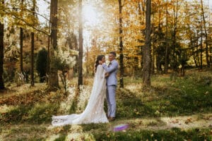 In einem sonnenbeschienenen Wald steht ein Paar im Hochzeitskleid inmitten herbstlicher Blätter. Der Bräutigam umarmt die Braut, die ein fließendes weißes Kleid trägt. Sonnenlicht fällt durch die Bäume und schafft eine warme und romantische Atmosphäre, perfekt für diesen wunderschönen Hochzeitsmoment.