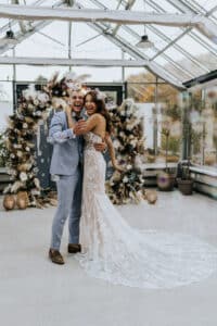 Ein fröhliches Paar feiert seine Hochzeit umarmt in einem sonnendurchfluteten Raum mit großen Fenstern. Die Braut trägt ein Spitzenkleid und der Bräutigam einen hellblauen Anzug. Hinter ihnen ergänzt ein dekorativer Blumenbogen in neutralen Tönen die festliche Atmosphäre ihres besonderen Tages.
