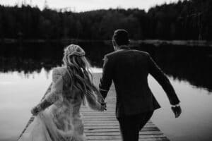 Ein Schwarzweißfoto zeigt ein Paar, das Hand in Hand zum Ende eines Holzstegs rennt. Mit Blick auf einen ruhigen, von Bäumen umgebenen See spiegelt die Szene den Charme einer Hochzeit wider; die Frau im Spitzenkleid ergänzt den Mann im Anzug, und sie verkörpern zeitlose Romantik.