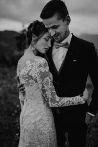 Ein Schwarzweißfoto fängt einen zärtlichen Moment der Hochzeit ein, bei dem sich Braut und Bräutigam im Freien umarmen. Die Braut in einem Spitzenkleid ruht mit ihrem Kopf auf der Brust des Bräutigams. Er trägt einen dunklen Anzug mit Fliege. Der unscharfe Hintergrund lässt auf eine ruhige, natürliche Umgebung schließen, perfekt für jede Hochzeit.