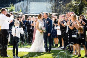 Braut und Bräutigam küssen sich bei ihrer Hochzeit, umgeben von jubelnden Gästen, die bei einer Hochzeit im Freien Seifenblasen pusten. Die Braut hält einen Blumenstrauß und der Bräutigam trägt einen Anzug mit einer Ansteckblume. Sonnenlicht fällt durch die Szene und verstärkt die festliche Atmosphäre ihres besonderen Tages.