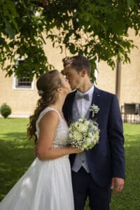 Als Teil ihrer Hochzeit küssen sich Braut und Bräutigam zärtlich unter einem Baum. Die Braut strahlt in ihrem weißen Kleid und hält einen Strauß weißer Rosen in den Händen, während der Bräutigam in seinem marineblauen Anzug mit hellgrauer Weste und Fliege schick aussieht, umgeben von üppigem Gras und einem charmanten beigen Gebäude.