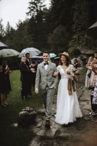 Während ihrer Hochzeit gehen Braut und Bräutigam fröhlich einen Weg im Freien entlang. Der Bräutigam trägt einen grauen Anzug, während die Braut in einem weißen Kleid mit Hut glänzt. Die Gäste stehen mit Regenschirmen inmitten üppigen Grüns und werden Zeugen dieses wunderschönen Heiratsmoments.