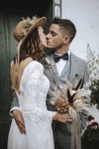 Braut und Bräutigam küssen sich vor einer grünen Tür und beginnen so ihre Hochzeitsreise. Die Braut in Spitzenkleid und Hut hält einen Strauß getrockneter Blumen in der Hand, während der Bräutigam einen grauen Anzug mit Fliege trägt. Die Szene strahlt rustikale Intimität aus.