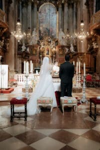 Braut und Bräutigam stehen in einer kunstvoll verzierten Kirche am Altar und genießen die Schönheit ihrer Hochzeit. Die Braut trägt ein weißes Kleid und einen Schleier, während der Bräutigam einen dunklen Anzug trägt. Kronleuchter, Statuen und aufwendige Dekorationen bereichern diesen unvergesslichen Moment der Hochzeit.