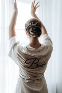 Eine Braut steht in der Magie ihrer Hochzeit versunken am Fenster, die Arme anmutig erhoben und ein dekoratives Haarteil tragend. Auf ihrem weißen Gewand ist stolz „Braut“ auf dem Rücken zu sehen, während das Sonnenlicht diesen bezaubernden Moment der Hochzeit sanft erhellt.