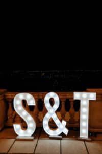 Große beleuchtete Buchstaben „S & T“ werden nachts auf einem Balkon angezeigt und schaffen eine romantische Szene, perfekt für eine Hochzeit, mit Blick auf eine Stadtlandschaft mit fernen Lichtern unter einem dunklen Himmel.