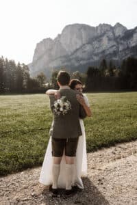 In einer malerischen Szene von Hochzeitsplanung umarmen sich Braut und Bräutigam auf einer Wiese mit Bergen im Hintergrund. Die Braut umklammert ihren Blumenstrauß, während der Bräutigam traditionelle Kleidung mit Kniestrümpfen trägt. Bäume rahmen das Feld elegant unter einem klaren Himmel ein.