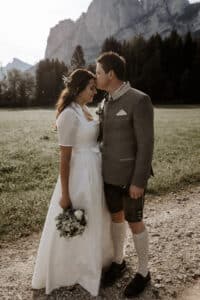 Ein Paar in traditioneller Kleidung teilt einen zärtlichen Moment im Freien und verkörpert damit die Essenz einer Hochzeit. Die Frau trägt ein weißes Kleid und hält einen Blumenstrauß, während der Mann in einer grauen Jacke sie auf die Stirn küsst. Sie stehen auf einem Schotterweg, der von malerischen Bergen und Bäumen gesäumt ist, und markieren den Beginn ihrer gemeinsamen Reise.