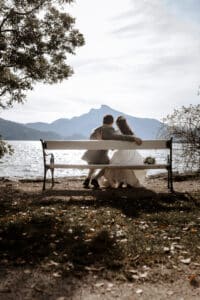 Ein Paar in Hochzeitskleidung sitzt auf einer Bank am See und blickt in der Ferne auf einen Berg. Die ruhige und romantische Atmosphäre ergänzt ihre Hochzeit, mit Bäumen, die die Szene einrahmen, und Sonnenlicht, das sich im Wasser spiegelt.