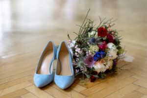 Ein Paar blaue High Heels steht neben einem farbenfrohen Blumenstrauß mit roten, weißen, rosa und blauen Blüten auf einem polierten Holzboden – die perfekte Ergänzung für jede Hochzeit oder elegante Hochzeitsatmosphäre.
