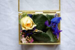 Eine dekorative Glasbox, perfekt für eine Hochzeit, mit einer gelben Rose, violetten Blumen, grünen Blättern und einem goldenen Verlobungsring, verziert mit kleinen Diamanten, alles wunderschön arrangiert, um Ihre Hochzeitsplanung zu verschönern.