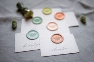 Sechs Karten mit eleganter Handschrift und farbigen Wachssiegeln in Grün, Gelb und Pfirsich werden auf einer Leinenoberfläche präsentiert und erwecken den Charme einer romantischen Hochzeit. Ein Wachssiegelstempel mit Holzgriff liegt daneben, perfekt, um Ihrer Hochzeitsplanung eine persönliche Note zu verleihen.