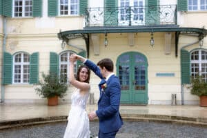 Eine Braut im weißen Kleid und ein Bräutigam im blauen Anzug tanzen fröhlich in einem Außenhof und verströmen die Essenz einer perfekten Hochzeit. Im Hintergrund ist ein gelbes Gebäude mit grünen Fensterläden und einer blaugrünen Tür zu sehen, was eine elegant festliche Atmosphäre schafft, die ideal für Hochzeiten ist.