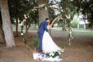 Ein Paar im Hochzeitskleid küsst sich unter einem runden Blumenbogen in einer Waldgegend, perfekt für ihre intime Hochzeit. Der Bräutigam trägt einen blauen Anzug und die Braut ein weißes Kleid. Blumen und Grünpflanzen schmücken den Bogen und unterstreichen die natürliche Umgebung.