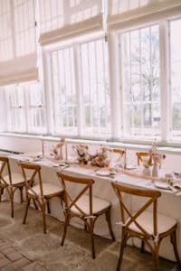 Ein sonnendurchfluteter Speisesaal, der sich perfekt für Hochzeitsfeiern eignet. Er verfügt über einen langen, elegant gedeckten Tisch mit weißer Tischwäsche, Blumenschmuck und Holzstühlen mit weißen Polstern. Große Fenster mit weißen Jalousien lassen natürliches Licht herein und schaffen eine ideale Kulisse für Hochzeitsmomente.