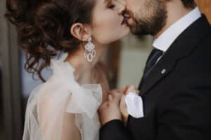 Während ihrer Hochzeit küsst sich ein Paar in formeller Kleidung im Haus romantisch. Die Frau trägt elegante Ohrringe und ein transparentes, gerüschtes Oberteil, während der Mann einen dunklen Anzug mit weißem Einstecktuch trägt. Sie halten zärtlich Händchen und schwelgen in der Freude des Heiratens.