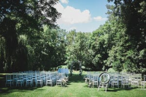 Eine Hochzeit im Freien mit Reihen weißer Stühle auf einem grasbewachsenen Gang, umgeben von üppigen grünen Bäumen. Elegante Blumenarrangements schmücken den Gang und rechts ist ein gespiegeltes Willkommensschild zu sehen. Der Himmel ist blau mit ein paar Wolken – eine perfekte Szene für eine Hochzeit in der Natur.