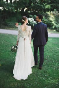Ein Paar in Hochzeitskleidung geht Hand in Hand auf einem Grasweg und verkörpert die Freude der Hochzeit. Die Braut trägt ein langes, elegantes Hochzeitskleid mit Spitzendetails und hält einen Blumenstrauß, während der Bräutigam einen dunklen Anzug trägt. Üppige grüne Bäume und ein Kiesweg runden diesen bildschönen Moment ab.