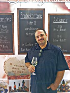 Ein Mann in einem dunklen Hemd hält ein Weinglas und steht vor einer Wand, die mit Weinangeboten und einem Schild auf Deutsch geschmückt ist. Die Umgebung lässt auf Kultiviertheit schließen, vielleicht perfekt für diejenigen, die eine Hochzeit planen oder schon weit fortgeschritten sind.