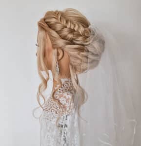 Eine Seitenansicht einer Braut mit einer eleganten geflochtenen Hochsteckfrisur zeigt ihren exquisiten Stil. Sie trägt ein Spitzenhochzeitskleid und einen langen Schleier und verkörpert damit die Essenz der Hochzeitsplanung. Zarte Ohrringe verleihen ihrem Look den perfekten Touch.