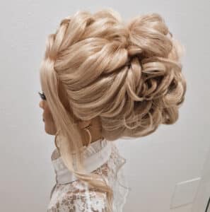 Eine Frau mit einer aufwendigen blonden Hochsteckfrisur mit Spirallocken und losen Strähnen, einem weißen Spitzentop und großen Creolen posiert vor einem schlichten weißen Hintergrund – perfekt geeignet für eine Hochzeit.