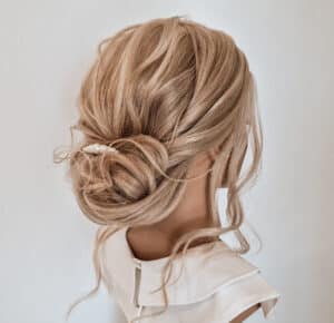 Das Haar einer Person ist in einer eleganten, lockeren Hochsteckfrisur mit weichen Locken und Strähnen gestylt, perfekt für eine Hochzeit. Das Haar ist mit kleinen Perlennadeln geschmückt und ergänzt das ärmellose weiße Oberteil mit Rüschenausschnitt.