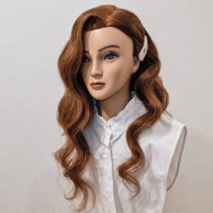 Ein Mannequinkopf mit langem, welligem, rotbraunem Haar, gestylt mit einer Perlenhaarspange, perfekt für die Eleganz einer Hochzeit. Er trägt eine ärmellose weiße Bluse mit Rüschenkragen vor einem schlichten hellgrauen Hintergrund.
