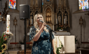 Eine Frau in einem blauen Blumenkleid singt in ein Mikrofon in einer Kirche und verleiht der Hochzeitszeremonie Magie. Im Hintergrund sind Buntglasfenster und kunstvolle Altardekorationen zu sehen, perfekt für alle, die heiraten und dabei eine elegante Note haben möchten.