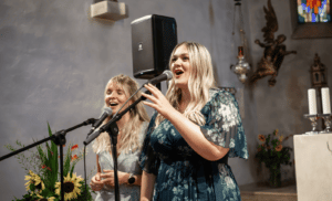 Zwei Frauen singen in Mikrofone in einer kirchlichen Umgebung, perfekt für jede Hochzeit. Eine trägt ein geblümtes Kleid und die andere ein helles Outfit. Das Dekor, ideal für Hochzeitsplanung, umfasst Kerzen, eine Engelsskulptur und ein Buntglasfenster mit Blumen und einem Lautsprecher in der Nähe.